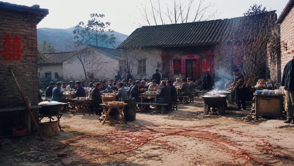 农村婚宴场景，八仙桌摆满大锅菜，人群围坐，喜字装饰，年代怀旧写实风格