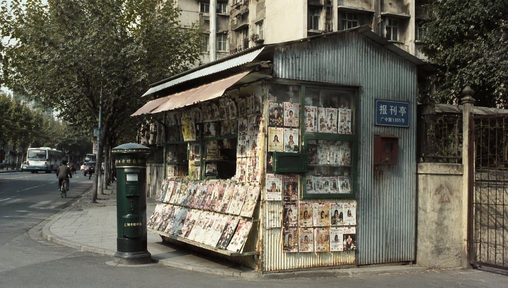 街角铁皮报刊亭，贴满旧杂志封面，旁边绿色邮筒，怀旧写实风格