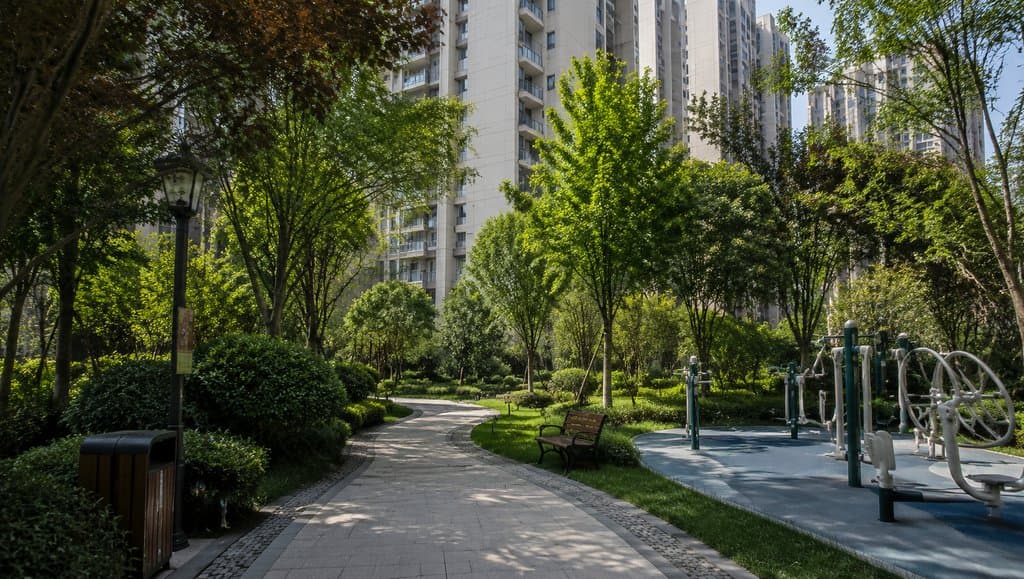 现代小区花园，绿树成荫的步道旁设有健身器材和长椅，背景为高层住宅楼，写实风格